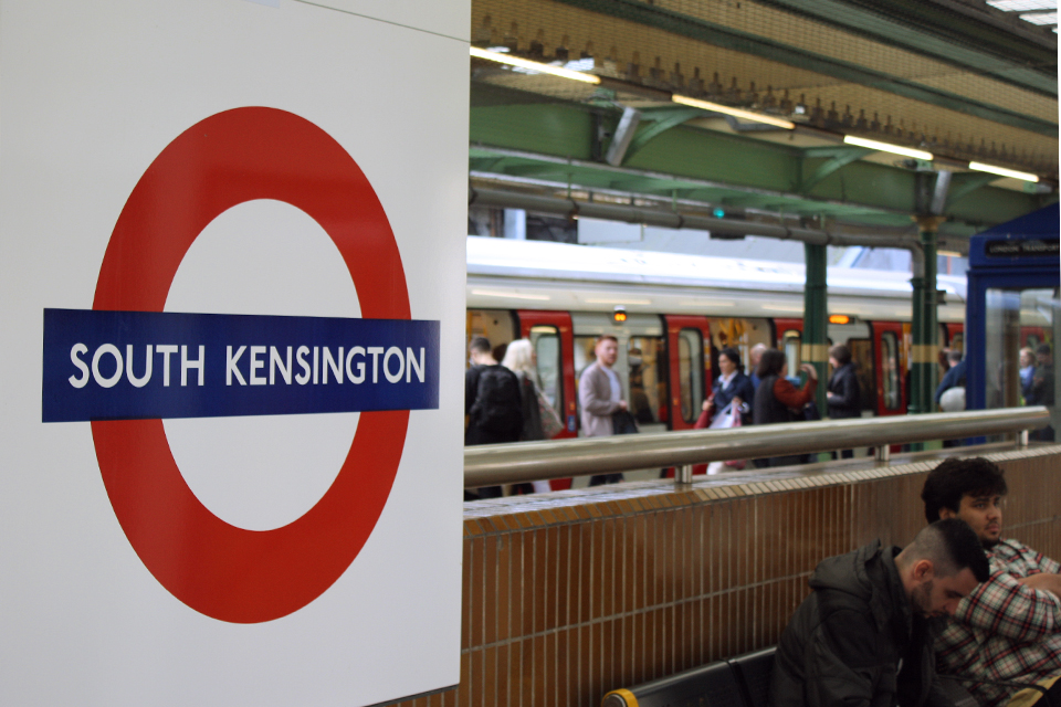 A close up of a Tube sign, stating South Kensington, with a tube symbol, with someone sitting at a seat, and a tube train behind them.